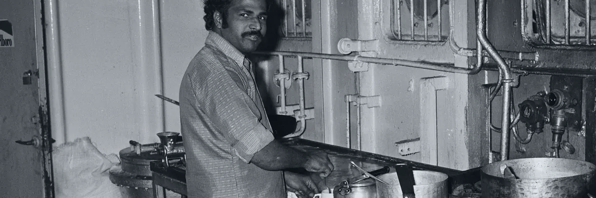 Black-and-white archive photo of a ship's cook at the stove in a small galley, pots on the range
