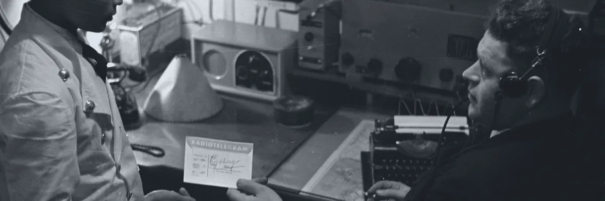 Black-and-white archive photo of the radio officer of a merchant ship at his set, headphones on, handing a message to a steward