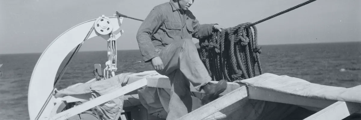 Black-and-white archive photo of a seaman working in a lifeboat swung out on its davits, the open sea behind